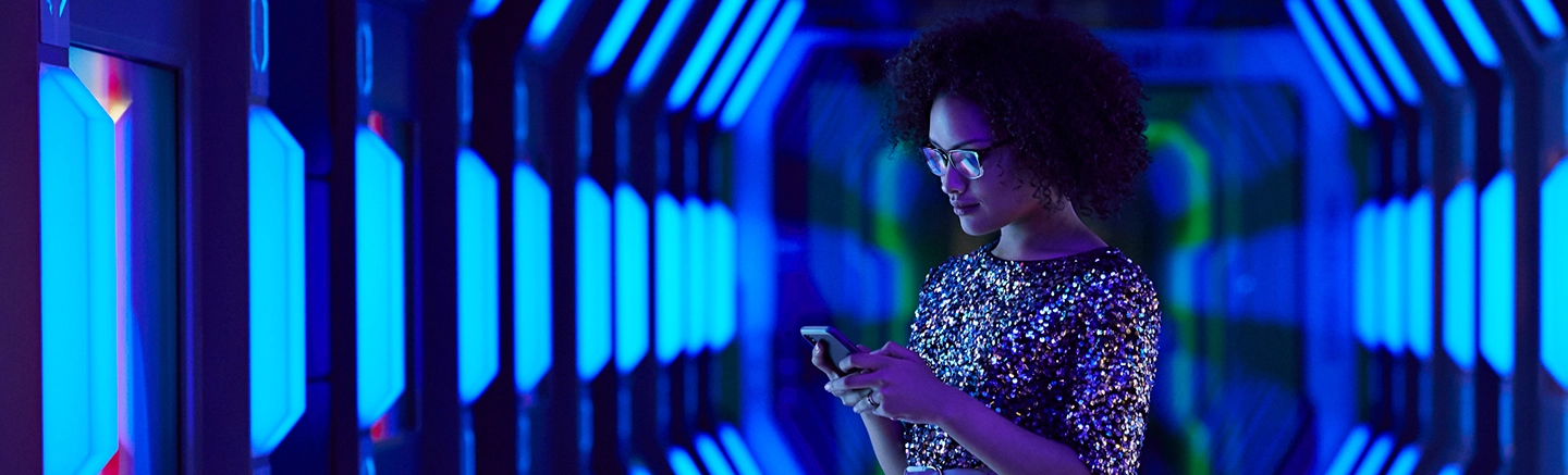 Hays Specialist Recruitment are the experts in Technology Industry Staffing. A woman with glasses and a sparkly shirt looks at her phone in an illuminated room with blue lights.