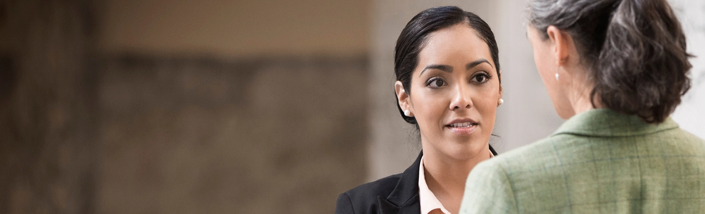A woman in a black business suit and cream-colored shirt speaks to another inside an office. The woman is wearing pearl-earrings and make-up.