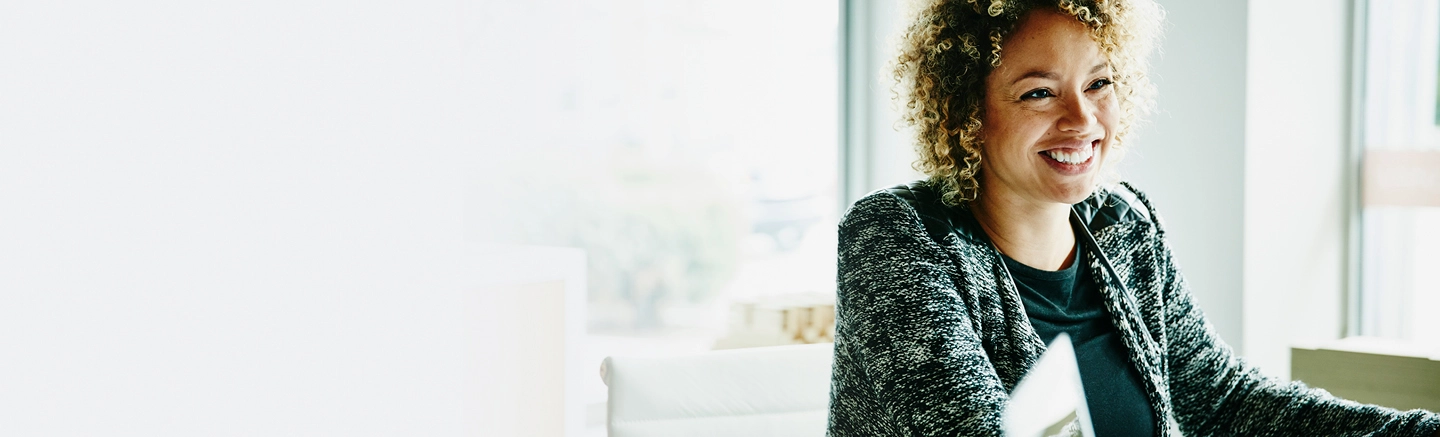 Black woman with curly hair smiling at colleague inside an office.