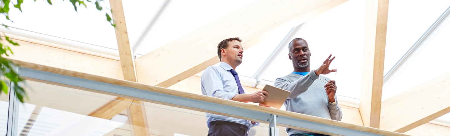 Two men in an office talk. One of them extends his hand outwards, while the second, wearing a blue shirt and a tie, looks on, holding a notepad.