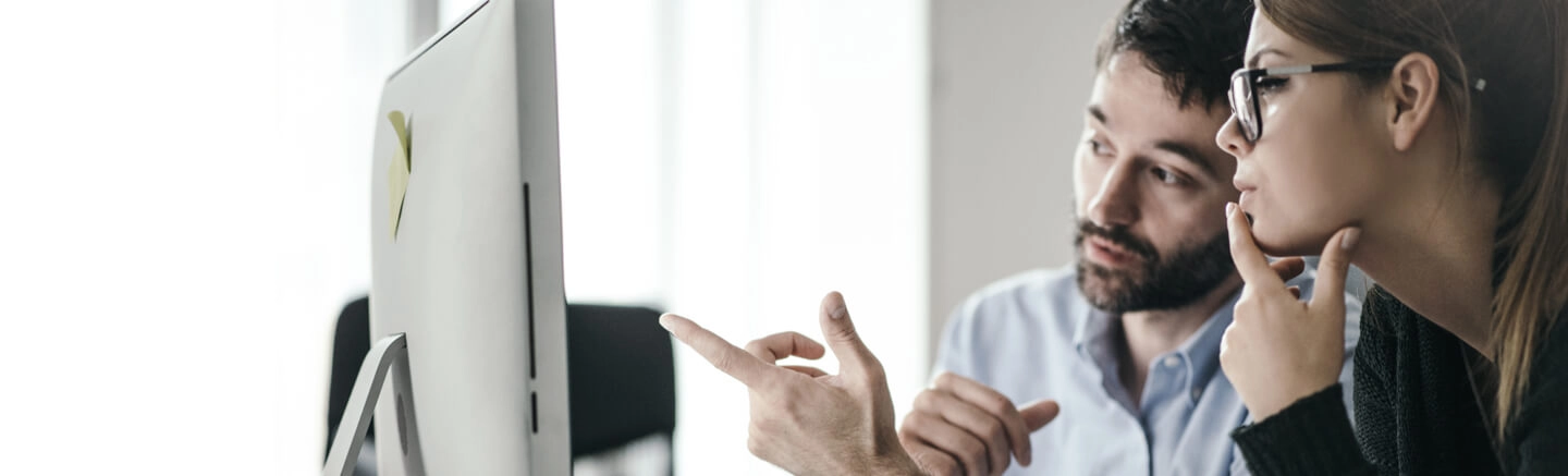 Two individuals with blurred faces seated at a desk in an office, looking at a computer screen. One person points at the screen while the other appears to be thinking, with a hand near their mouth.