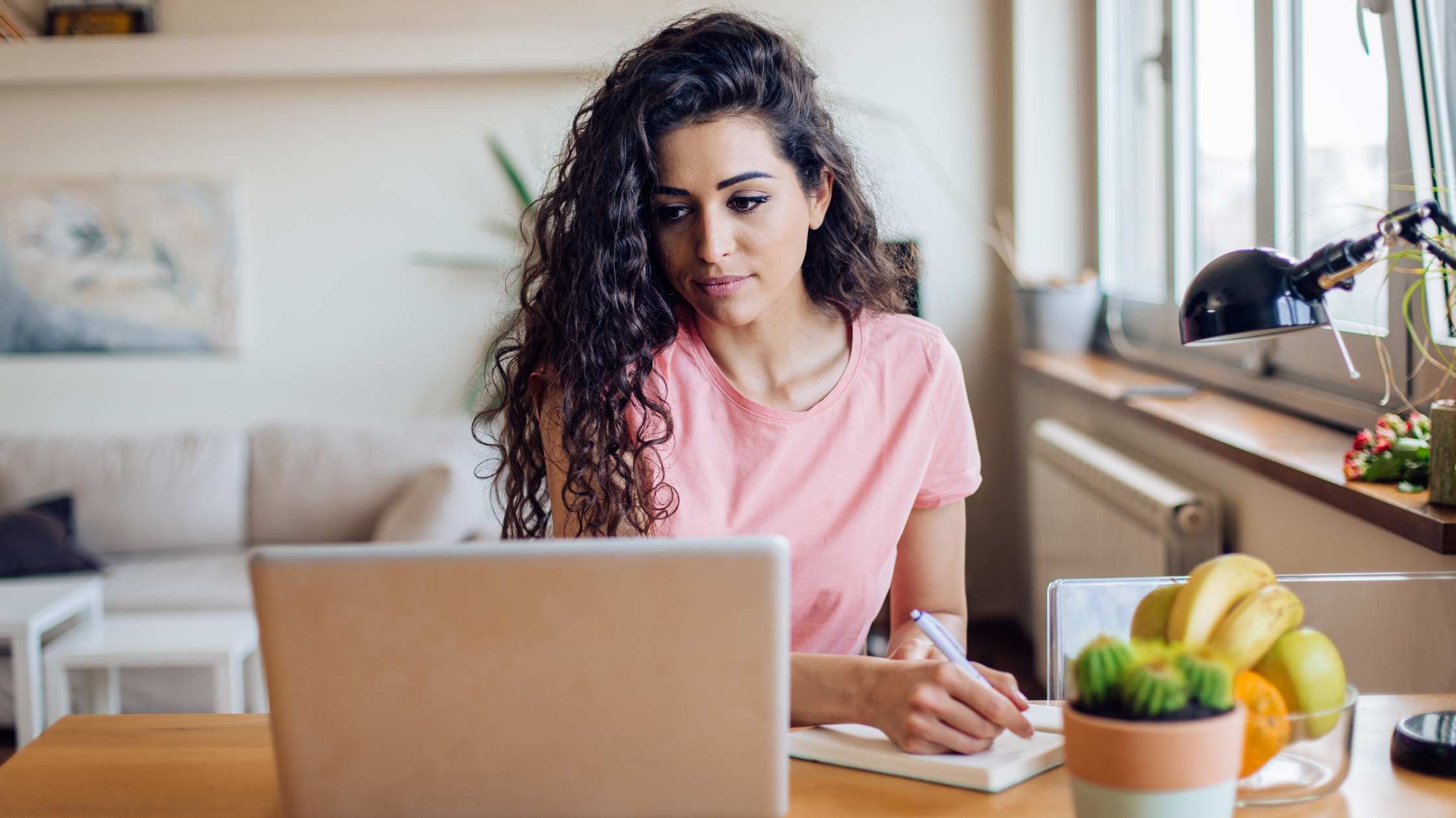 A woman in a pink shirt makes some notes on a notebook as she looks into a laptop. She is in in a living room, working from home.