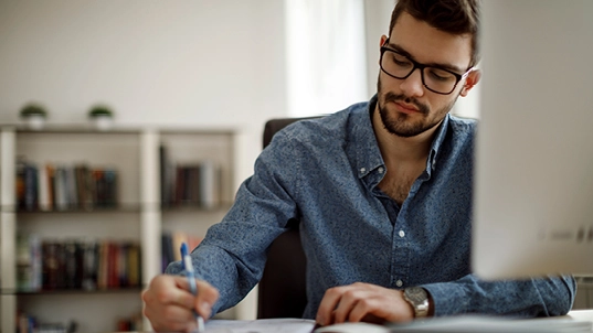 A man writes on a notebook. He wears a blue shirt and glasses and is sitting with a monitor in on the right hand side.
