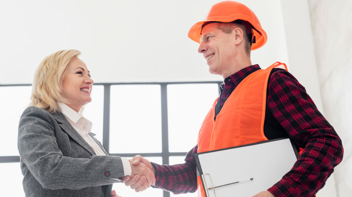 How to Build Relationships as a Contractor: A blond woman in a grey suit shakes hands with a man on a flannel shirt and bright orange Construction gear. Both are smiling at each other.