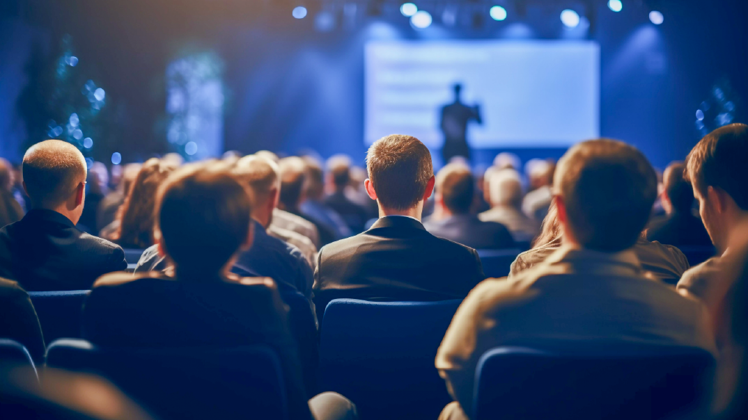 A blue-tinged image of a group of people wearing suits and attending a conference. On the background, blurred, is a person presenting.