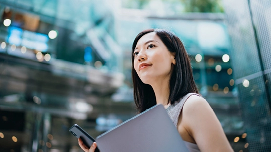 A woman holds a black phone and a binder. She's surrounded by buildings.