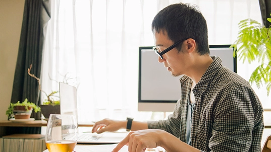 A man works from home in his laptop, next to him there's a glass with a golden beverage and behind him there is a computer monitor.