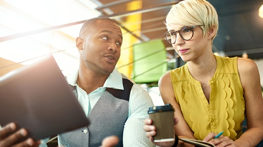 A man holding a tablet looks a a woman with a yellow blouse and glasses. The woman is holding a coffee cup and a pen and notepad.