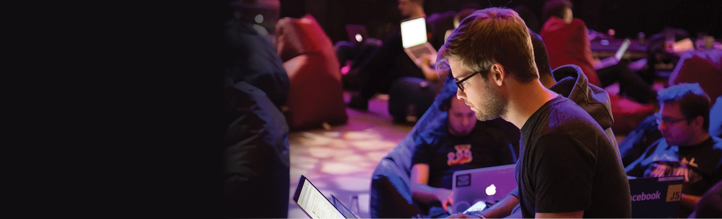 A group of people sitting on bean bags and working on laptops in a dimly lit room, suggesting a casual or collaborative setting like a hackathon or tech event.