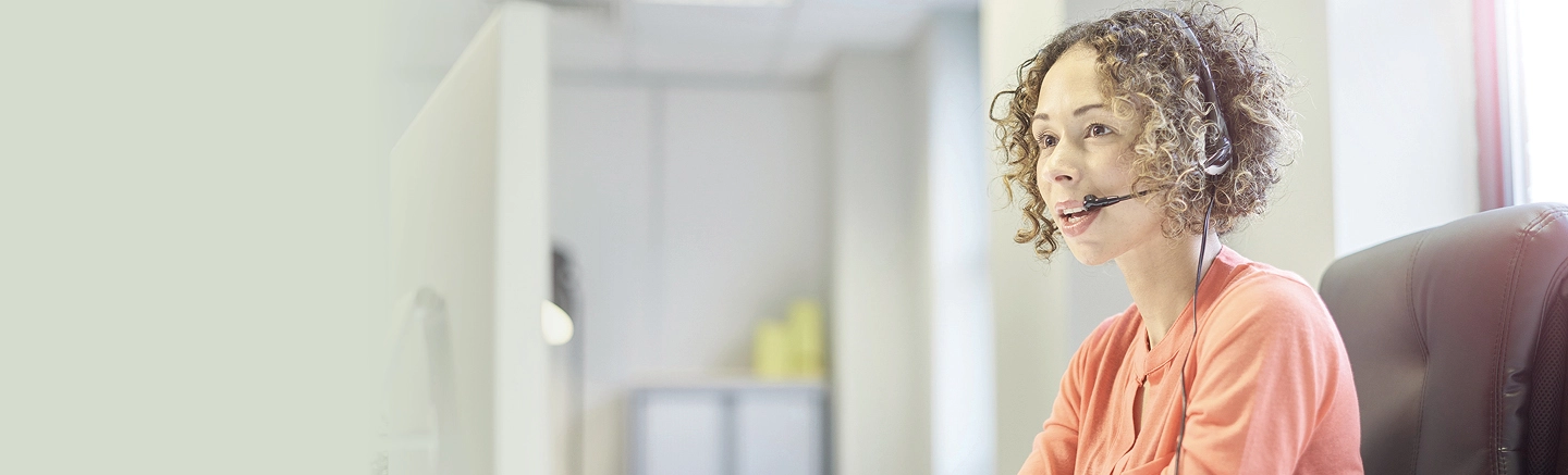 Person with curly hair wearing a coral-colored top, seated in an office chair and facing a computer screen. Their face is blurred for privacy. Office shelves with various items are visible in the background.