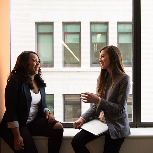 Two coworkers talking by office window with tablet.
