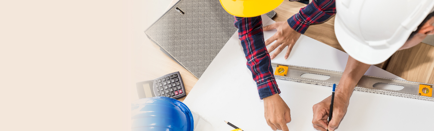 Construction workers drawing on a table. They're wearing yellow and white hardhats and one of them a blue and red flannel shirt. On the table is also a calculator and a blue hardhat.
