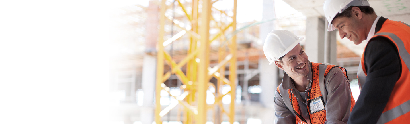 Two senior construction workers looking at plans on a high-rise site. Both are wearing white hard-hats and bright orange construction vests.